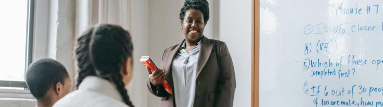 Teacher smiling at students, standing beside whiteboard with questions written on it